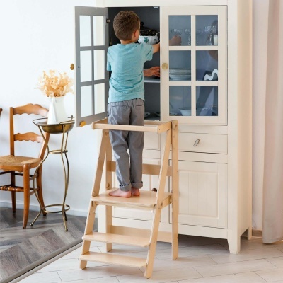 Criança usando escadote de madeira para alcançar móvel branco com portas de vidro