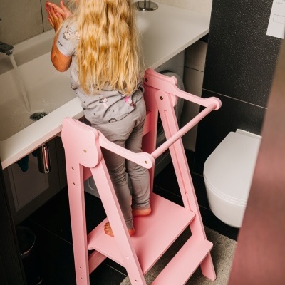 Criança loira a lavar as mãos num banheiro usando um banquinho rosa de madeira