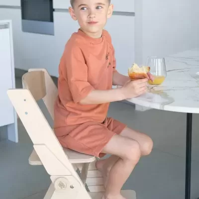 Criança sentada em cadeira de madeira clara junto a mesa branca com copo de sumo