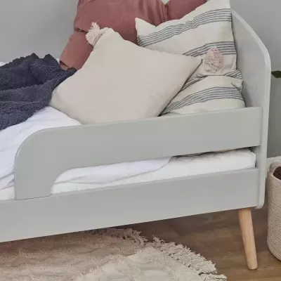 Cama cinza claro com colchão branco, almofadas bege, rosa e cinza, tapete bege e cesta de palha junto ao chão