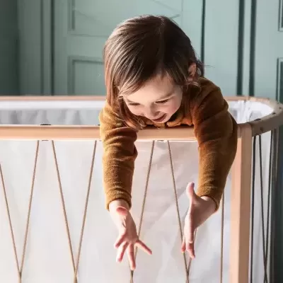 Berço de madeira clara com criança loira esticando os braços para fora