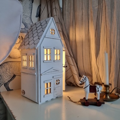 Casa decorativa branca com luz interior, cavalo de brinquedo de madeira e velas em mesa branca com cortinas bejes