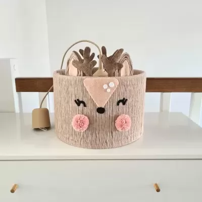 Iluminação decorativa em forma de cabeça de veado rosa claro com detalhes castanhos e pompons cor de rosa sobre móvel branco