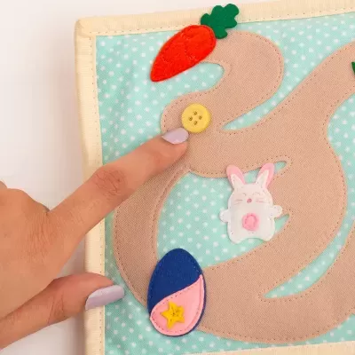 Jogo sensorial em tecido com percurso bege, botão amarelo, cenoura laranja, ovo azul e rosa, coelho branco e rosa