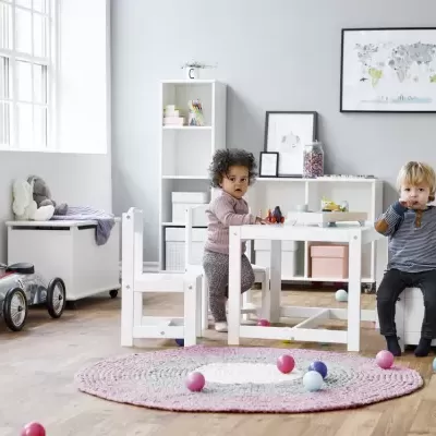 Quarto infantil com mesa e cadeiras brancas, tapete rosa e crianças brincando