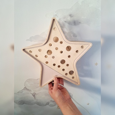 Candeeiro de parede em forma de estrela de madeira clara com furos circulares, com mão a segurá-lo e papel de parede de nuvens ao fundo