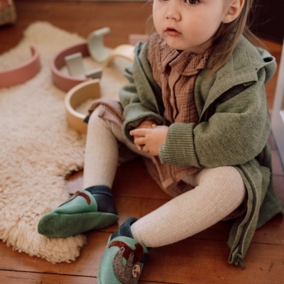 Bebé com sapatos verde de pele com aplique, vestido castanho e casaco verde sentada no chão de madeira perto de tapete branco felpudo.