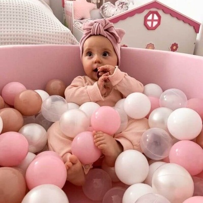 Bebé numa piscina de bolas rosa e branca, com roupa rosa e turbante, num quarto com cama branca e casinha de bonecas rosa e branca.