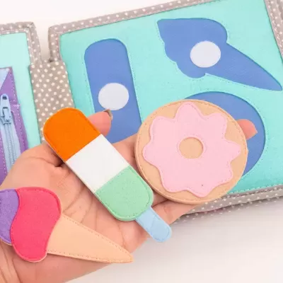 Brinquedo educativo de tecido com formas de gelado e bolacha coloridos em fundo azul e cinzento às bolinhas