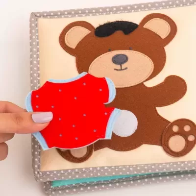 Livro de atividades em tecido com urso castanho e peça de roupa vermelha para vestir o urso