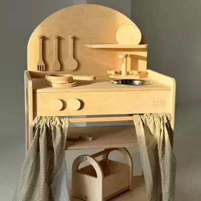 Cozinha de brincar infantil em madeira clara com utensílios, pia metálica e cortinas xadrez.