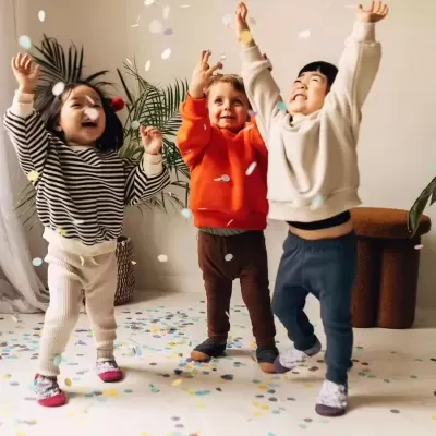Três crianças brincando com confetes dentro de casa vestindo roupas de inverno.