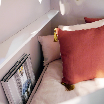 Almofadas em linho cor tijolo com tassel dourado numa cama com roupa bege e livros encostados na parede branca