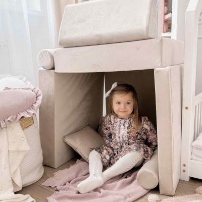 Menina sentada dentro de cabana feita com almofadas bege, cobertor cor de rosa no chão e decoração neutra ao redor