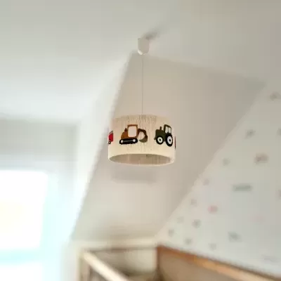 Lâmpada de teto com abajur branco e desenhos de veículos coloridos acima de berço de madeira