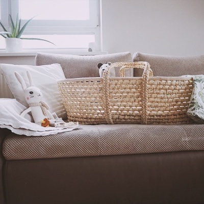 Cesto trançado em fibra natural com alças num sofá castanho claro com almofadas e peluche.