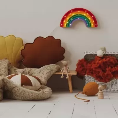 Almofadas em forma de concha, manta bege, bola, cesta com manta vermelha e arco-íris decorativo numa parede branca.