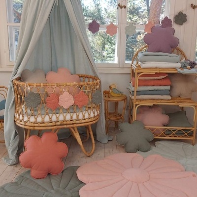 Berço de bebé em vime com almofadas florais e dossel cinzento num quarto claro com janela e tapetes florais no chão.