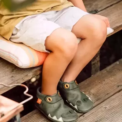 Criança com sapatilhas verdes de pele com design de rosto de animal sentada num banco de madeira.