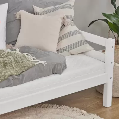 Cama de criança branca com colchão e almofadas decorativas ao lado de planta e tapete