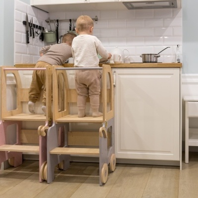 Duas crianças em escadotes em cozinha com armários brancos e parede de azulejos