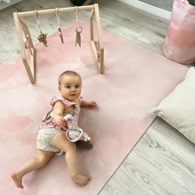 Criança deitada num tapete rosa claro com brinquedos pendurados num suporte de madeira.