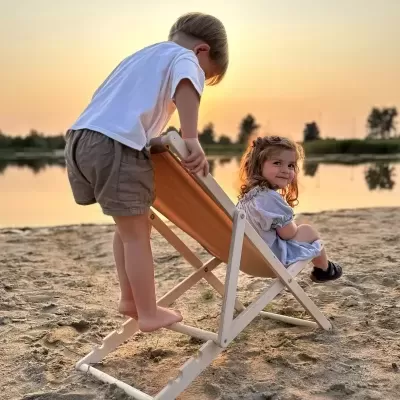 Cadeira de praia infantil de madeira com assento laranja na areia com duas crianças ao pôr do sol