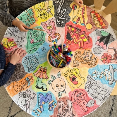 Mesa redonda colorida com desenhos de letras e canetas no centro, crianças pincelando os desenhos.