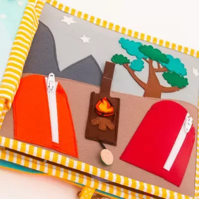 Livro infantil de tecido com tendas, fogueira e árvore feitas em feltro