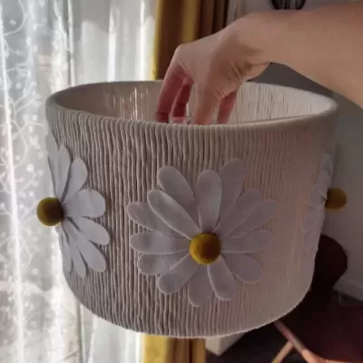 Abajur com cúpula bege texturizada com flores brancas e centro amarelo, segurado por mão