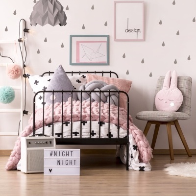 Quarto infantil com cama de grades preta, manta rosa, almofadas, candeeiro cinzento e decoração em tons claros