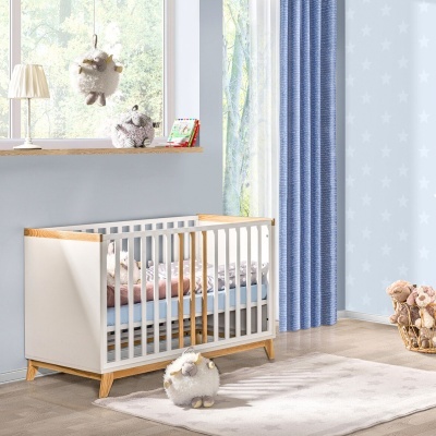 Berço de bebé em madeira clara e branca com manta rosa e colchão azul num quarto com tapete bege, cortinas azuis, e peluches.