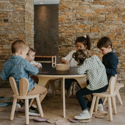 Crianças sentadas à mesa em cadeiras e mesa de madeira clara com parede de pedra ao fundo