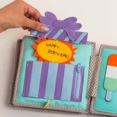Livro têxtil infantil com presente lilás e etiqueta amarela com 'HAPPY BIRTHDAY!'
