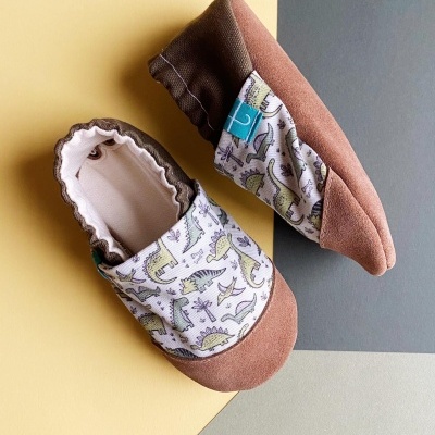 Sapatinhos de bebé com padrão de dinossauros em tecido castanho claro e branco.