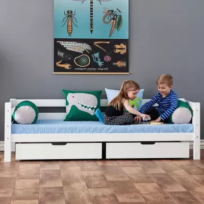 Cama infantil branca com gavetas, colchão azul e almofadas de dinossauro com duas crianças a brincar
