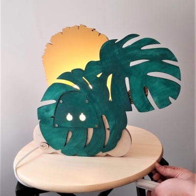 Candeeiro de mesa com folhas verdes Monstera e luz amarela num suporte de madeira clara sobre mesa redonda clara com pernas pretas.