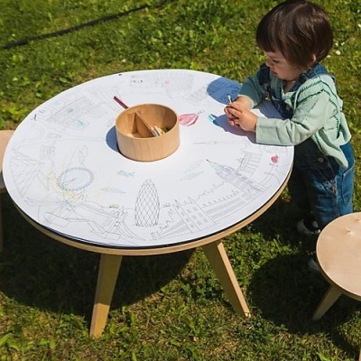 Mesa redonda branca com desenhos para colorir, criança a pintar, mesa com pernas de madeira clara na relva.