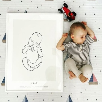 Pôster de desenho de bebé em moldura branca junto a bebé real adormecido sobre lençol decorado com barcos e estrelas