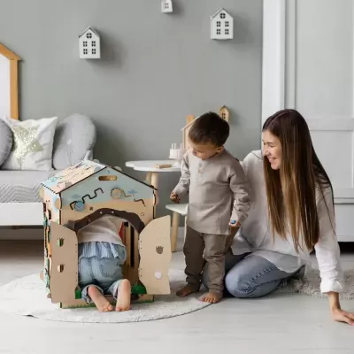Crianças brincando com casa de papelão decorada dentro de sala com mãe e decoração cinzenta