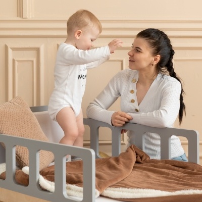 Berço cinza com roupa de cama castanha e almofada rosa, bebê e mulher ao lado