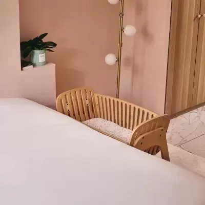 Berço de madeira clara junto à cama num quarto decorado com parede rosa, candeeiro dourado e planta em vaso verde