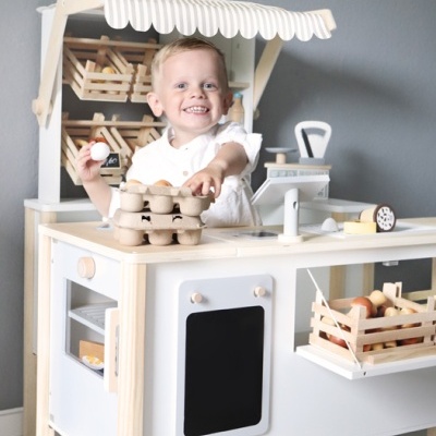 Criança brincando com banca de jogo de cozinha em madeira clara e branca com acessórios de supermercado.