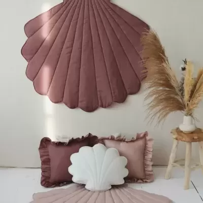 Tapete e almofadas formato concha em tons rosa e branco com textura acolchoada, arranjo de plantas secas e banco de madeira