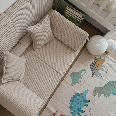 Sofá beige de veludo cotelê com almofadas e tapete infantil de dinossauros sobre chão de madeira