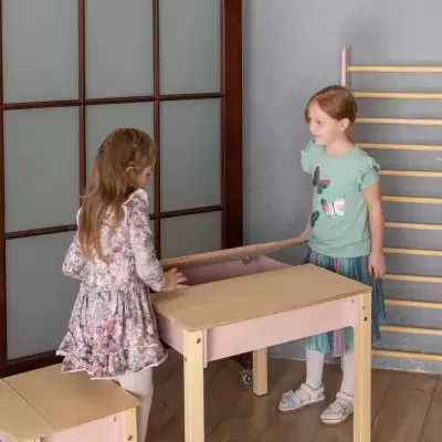 Duas meninas a brincar com mesa e bancos infantis de madeira clara e rosa num quarto com parede cinza.