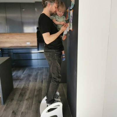 Homem em pé num banquinho branco com criança, numa cozinha moderna com paredes branca e preta onde estão desenhos coloridos colados.