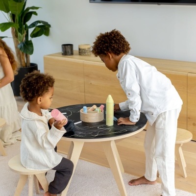 Crianças a desenhar num tampo de mesa redonda preto com giz de cera colorido, em ambiente interior com móvel de madeira clara e planta verde