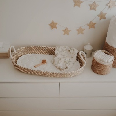 Cesto de vime com cobertores e acessórios de bebê sobre uma cômoda branca, com decoração de estrelas penduradas na parede.