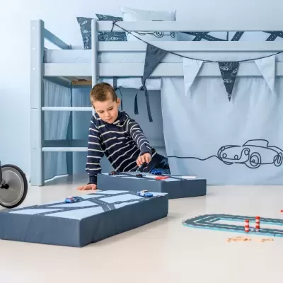 Cama alta de criança azul claro com cortinas e bandeirolas decorativas e menino a brincar com carros de brinquedo no chão